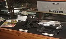 Une vitrine de expose des accessoires utilisés pendant le tournage du film Men in Black 2. À gauche, un magazine factice avec en grosse lettre « Man awakens from 35-year coma », au centre un pistolet chromé minuscule sur un coussin noir et à droite un pistolet de science fiction ressemblant à un pistolet normal surmonté d'un second canon, lui aussi sur coussin noir. Une affiche du film se reflète sur le verre.