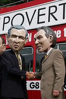 Photographie couleur de deux personnes qui portent les masques de George W. Bush et Tony Blair et se sèrent la main.