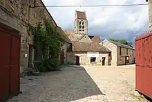 L'ancienne cour de l'église a été reconvertie en zone d'activité artisanale et de bureaux. Au premier plan, portail en fer ouvert ; à gauche, vieux bâtiment, fenêtres modernes entourées de glycine ; en face bâtiment ancien d'un étage, avec église en arrière-plan, à gauche bâtiment en biais