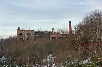 Photo hivernale montrant deux bâtiments en ruine sans toiture et envahi par la végétation dont l'un éventrée ainsi qu'une cheminée d'usine tronquée.