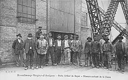 Photographie noir et blanc montrant un groupe de mineurs au visage noirci posant devant un bâtiment en briques.