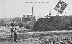 Photo en noir et blanc d'un ensemble de bâtiments industriels typés 1900 avec deux grands chevalements (tour avec bigue) métalliques et cheminée d'usine.