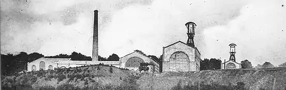 Photo noir et blanc, un ensemble de bâtiments industriels typés 1900 avec deux grands chevalements (tour avec bigue) métalliques et cheminée d'usine.