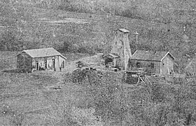 Photo noir et blanc d'un chevalement en bois encadré par deux petits bâtiments maçonnés.