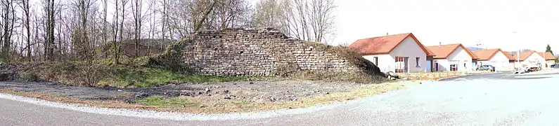 Vue panoramique d'un vestige de mur, sur la gauche se trouve des arbres et la lampisterie, sur la droite se trouve des maisons neuves de lotissement.