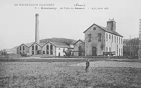 Photo noir et blanc montrant un ensemble de bâtiments industrielles dont un très haut abritant le chevalement, ce dernier dépasse surmonté d'un clocher en bois. Sur la gauche se trouve une grande cheminée.