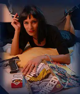 Une femme brune allongée sur le ventre les jambes à demi-repliées et fumant une cigarette, avec un pistolet et un magazine posé devant elle, parodiant Uma Thurman sur l'affiche du film.