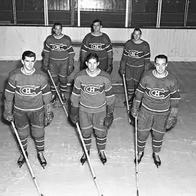 Photographie en noir et blanc de six joueurs des Canadiens de Montréal en pied. Les joueurs sont sur la glace répartis en deux rangs et près à jouer au hockey sur glace