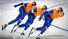 Trois patineurs en combinaison orange, l'un à la suite de l'autre, en fin de virage.