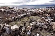 Photographie en couleurs de blocs de pierre non taillés installés à terre sous forme rectangulaire, une étendue herbue au centre, des eaux maritimes encadrées de reliefs montagneux visibles en arrière-plan.