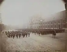 Photo en noir et blanc de militaires en uniforme en formation incluant une fanfare à la gauche de la formation principale et un militaire monté à cheval se tenant devant celle-ci