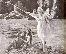 À la gauche de la photo, un chevalier qui semble blessé est supporté par une femme, les deux étant assis par terre. À la droite, une fée en position debout brandit une baguette.