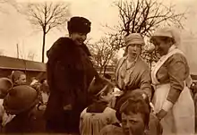 à gauche Henriette vêtue d'un manteau et d'une toque en fourrure souriante regarde un enfant qui reçoit un présent de la reine Élisabeth coiffée d'un bandeau aux côtés d'une infirmière souriante de profil