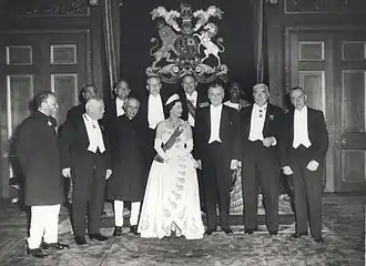 Élisabeth II en robe du soir et portant une tiare est entourée de onze hommes en tenue de soirée ou en costume national.