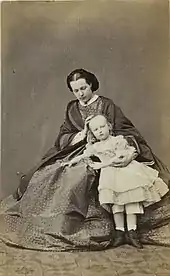 La reine assise et portant une robe à crinolines est légèrement inclinée, le visage grave, vers sa fille debout en robe claire, le regard fixant l'objectif du photographe.