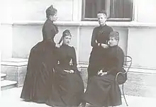 photographie de quatre femmes en deuil, dont deux sont assises et deux debout posant devant le sous-bassement d'un bâtiment en pierre claire.