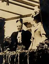 À gauche la reine, portant une courte veste, un col à jabots et les cheveux coiffés en arrière pose face à l'objectif, à droite Clémentine, de profil, porte une longue tenue claire et est coiffée d'un chapeau à fleurs