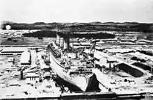 A large ship inside of a dry dock. The dry dock is surrounded by industrial buildings and hills are visible in the background