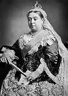 Photographie en noir et blanc de la reine Victoria coiffée d'un voile en dentelle surmonté d'une petite couronne, elle tient en main un éventail fermé.