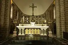 Photographie d'un autel en marbre de style néo-roman, surmonté d'une grande croix, avec des petits chandeliers, et deux grands sur les côtés