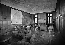 Un salon aux murs et au plafond couverts de boiseries. Une dizaine de fauteuils entourent deux tables basses et une grande carte de la Scandinavie est affichée face à un bureau.