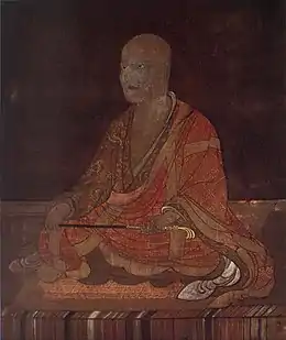 Moine bouddhique assis en tailleur, sceptre entre les mains. Vêtements orangé, brun et jaune sur fond brun foncé.