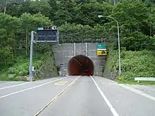 .Photo couleur de l'entrée d'un tunnel entouré de verdure. Une route à deux voies au premier plan.