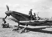 Trois hommes s'affairant autour d'un avion. L'un est debout sur l'aile et regarde vers le photographe, un autre regarde dans le cockpit tandis qu'un troisième s'éloigne en tirant un chariot.