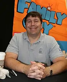 Homme aux cheveux bruns et souriant qui se tient derrière une table d’interview.