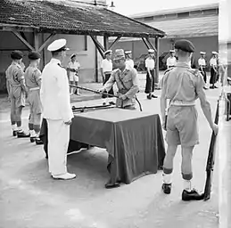 Un homme asiatique déposant des armes sur une table derrière laquelle se tiennent, debout, un militaire occidental et d'autres soldats.