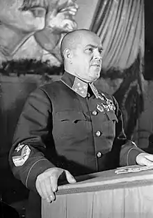 Photo noir et blanc d'un homme en uniforme faisant un discours à un podium, les profils de Staline et de Lénine derrière.