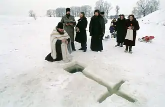 Un pope à genou devant une croix creusée dans la glace d'une rivière gelée. Une dizaine d'assistants, dont un homme tenant une bible ouverte et une femme avec des bougie allumées.