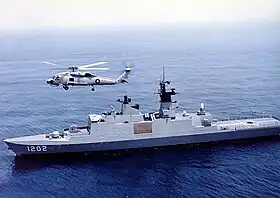 Un navire de grande taille, gris, avec un hélicoptère volant au-dessus.