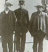 Leovigildo Galarza (vêtu d'un pantalon, d'une veste et d'un chapeau) et Jesús Carvajal (vêtu d'un pantalon et d'un poncho) accompagnés d'un geôlier en uniforme.