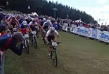  Peloton de plusieurs dizaines de coureurs à VTT avec en arrière-plan un public derrière une barrière.