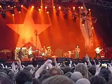 Photo d'un concert de Rage Against The Machine dans laquelle on voit au premier-plan, de dos, les têtes des spectateurs et des bras levés, et en arrière-plan, la scène dont l'éclairage donne un effet de couleur orangée sur laquelle les membres du groupe sont en train de jouer ; le fond de la scène est constitué d'une toile sur laquelle est dessinée une très grande étoile blanche.