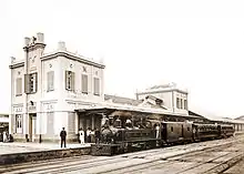 Photographie d'une locomotive à vapeur avec des wagons de passagers devant les grands bâtiments de la gare