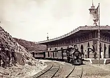 Photographie d'une locomotive à vapeur et ses wagons sur une voie en courbe et le bâtiment de la gare