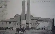 Carte postale ancienne en noir et blanc montrant la fosse n° 2 des mines de Vicoigne à Raismes, avant la Première Guerre mondiale. Le chevalement ne se détache pas vraiment du bâtiment, et qui plus est, il y a deux cheminées massives devant. Plusieurs chevaux sont visibles.