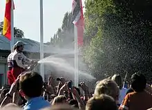 Sébastien Loeb et Daniel Elena en combinaison automobile, vus de profil sur une estrade, sabrant des bouteilles de champagne entourés par une foule de spectateurs.