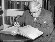 Un homme aux cheveux blanc, costume trois pièces, lunettes, oreillette d'un appareil auditif, en train d'écrire de son stylo plume, sur un gros ouvrage relié