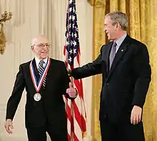 Un homme souriant en costume noir porte une médaille autour du cou, et un homme souriant en costume noir le congratule, avec sa main sur son épaule.
