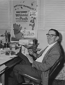 Photo en noir et blanc de l'animateur Ralph Wright portant des lunettes noires assis à son bureau en 1968 avec une affiche du film Winnie l'ourson et l'Arbre à miel accrochée au mur.