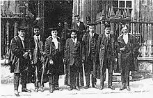Photographie noir et blanc d'un groupe de sept hommes, posant, de face, en toge noire de professeur d'université, devant l'entrée d'un bâtiment.