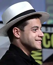 Un homme de profil, souriant, aux cheveux courts et bruns et portant un chapeau noir et blanc.