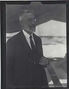 Photo en noir et blanc d'un homme d'âge mur, portant lunettes, cheveux et barbe blancs, debout, tourné vers la droite, habillé en costume-cravate.