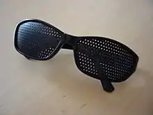 Paire de lunettes en plastique noir, aux branches repliées, dont les verres sont remplacés par une plaque de plastique noir percée sur toute sa surface de plusieurs séries de trous de faible diamètre.