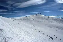 Sommet d'une piste de ski, avec une remontée mécanique et de nombreux skieurs éparpillés.