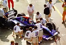 Photo en plongée sur une Formule 1 violette lors de sa sortie du garage, entourée de mécaniciens.