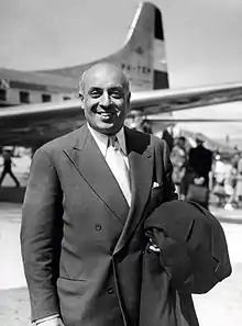 Photographie en noir et blanc d'un homme en costume sur le tarmac d'un aéroport, un avion en arrière-plan.